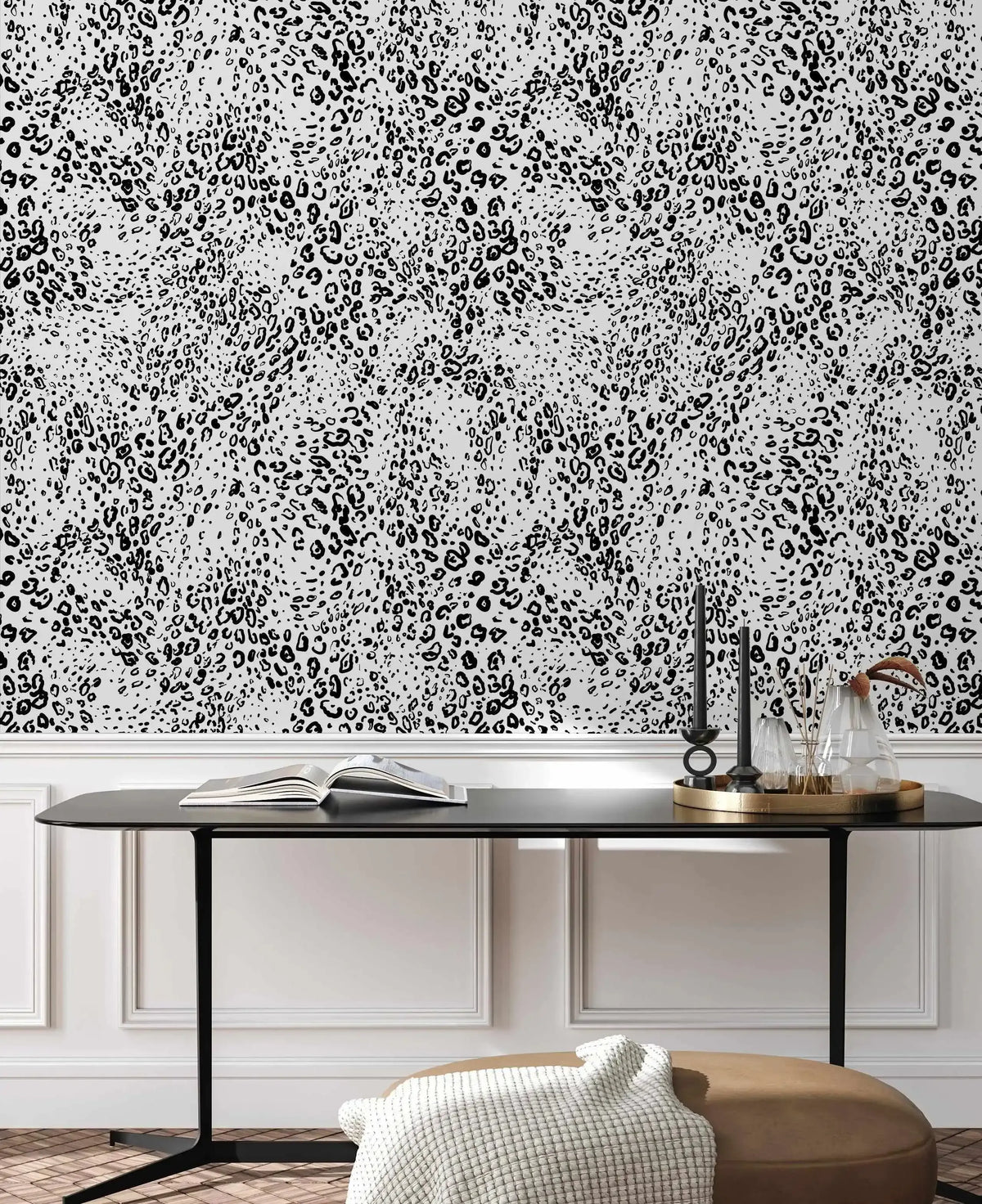 Dining Room:
Animal print wallpaper featuring a repeating pattern of black leopard spots and small dots on a light grey background, displayed above white wainscoting in a dining room. A black dining table with a gold tray holding black candles and glass vases sits in front of the wallpaper. A cream textured throw is draped over a brown ottoman on the wooden floor.