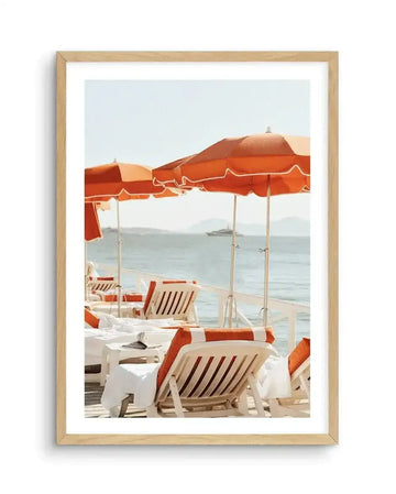 Framed art print in natural wood frame with white border showing a vintage beach scene. The photograph depicts a seaside terrace with rust-orange umbrellas and striped loungers in cream and terracotta tones. White-clothed tables are arranged on sandy beach in foreground, with calm turquoise ocean and distant mountains visible beyond. Clear sky transitions from white to pale blue. The composition captures a serene, classic Mediterranean resort aesthetic in soft, warm tones.