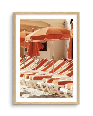 Framed art print depicting a Mediterranean beach scene with rust-red and cream striped lounge chairs and matching umbrellas casting diagonal shadows on sandy ground. Cream-colored buildings with arched architecture visible in soft, warm background tones. Natural wood frame with white border. Watercolor or gouache painting style with muted terracotta, beige, and cream color palette evoking a vintage 1970s aesthetic.