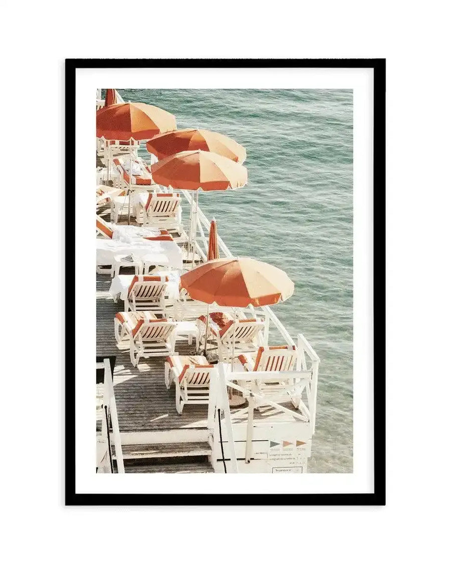 Framed art print showing a vintage color photograph of a ship's deck with four rust-orange and cream striped umbrellas shading white wooden deck chairs arranged in a row along a wooden railing. Turquoise-green ocean water fills the background. The photograph has a warm, nostalgic 1970s aesthetic. The print is displayed in a black frame with a white border.