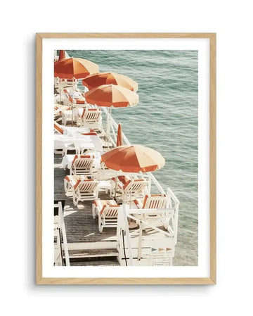 Framed art print featuring a vintage photograph from 1972 showing a ship's deck lined with white lounge chairs and orange-and-cream striped umbrellas overlooking turquoise-green ocean water. The photograph has warm, saturated colors with a nostalgic quality. Displayed in a natural wood frame with white border, evoking mid-century leisure and travel aesthetics.