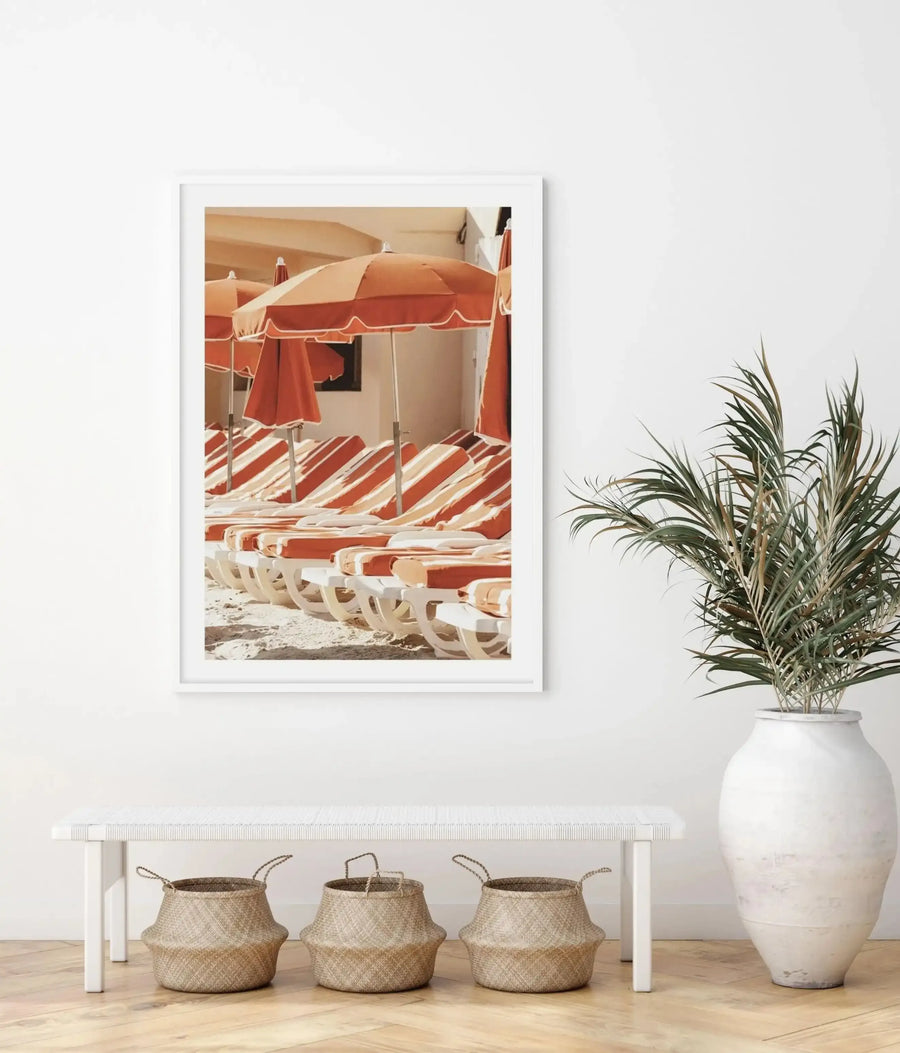 White framed art print hung on a light gray wall above a white wooden bench. The print depicts a sunny beach scene with red and white striped umbrellas, matching striped loungers, a red changing cabin, and neutral sand tones in a vintage illustration style. Below the framed print sits the white bench with three woven natural fiber baskets underneath, and a white ceramic vase with dried palm fronds to the right. Warm, coastal aesthetic with cream, coral, and tan color palette.