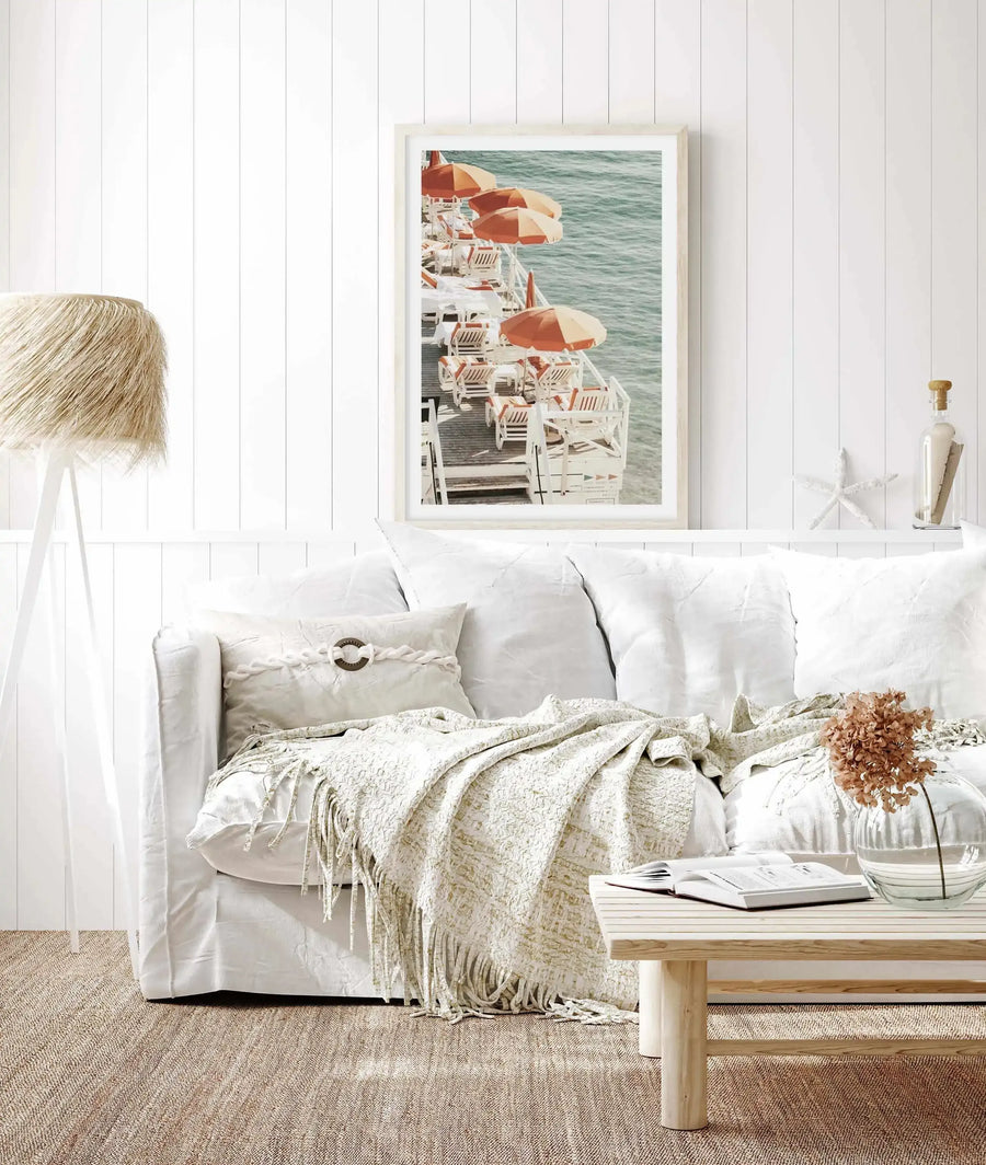 Framed art print displayed on a white shiplap wall above a white linen sofa. The photograph shows a vintage coastal scene from 1972 featuring white wooden deck chairs arranged on a ship's deck, with coral and cream striped umbrellas shading the seating area. Turquoise water is visible in the background. The print has a cream-colored frame with white border. The room styling includes neutral tones with natural wood accents, a woven throw blanket, and coastal decorative objects on a shelf beside the artwork.