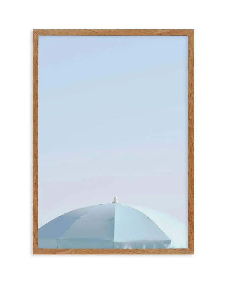 Framed photograph of a light blue parasol against a pale blue sky, with natural wood frame and white border. The parasol is cropped at the bottom, showing the top half of its dome and a small white finial at its peak. The sky is clear and bright, filling the upper two-thirds of the composition.