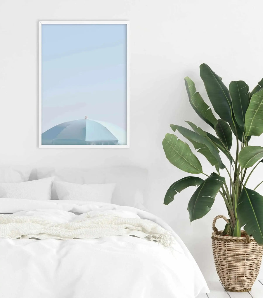 Lifestyle shot of a minimalist bedroom featuring an art print of an aqua parasol against a pale blue sky. The print is displayed in a white frame with a white border above a bed with white bedding and a cream textured throw. To the right, a large potted plant with dark green leaves sits in a woven basket.