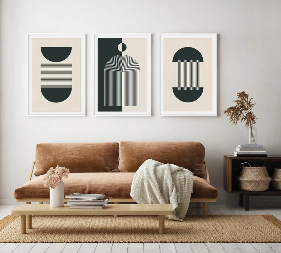 Three framed art prints displayed above a brown sofa in a minimalist living room. The prints feature abstract geometric shapes and lines in dark teal and cream, with white frames and white borders. The room includes a light wood coffee table with a white vase of dried flowers, a woven rug, and a dark wood side table with books and dried pampas grass.