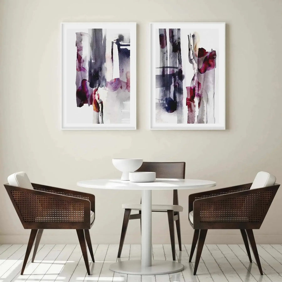 Lifestyle shot of a dining area with two framed abstract art prints displayed above a round white table with two white bowls. The prints feature abstract vertical brushstrokes in shades of deep plum, dark grey, and muted purple, with hints of red and black, on a light cream background. The dining area includes two dark wood chairs with woven cane backs and white seat cushions, and a third chair partially visible behind the table. The floor is white wood with visible planks.