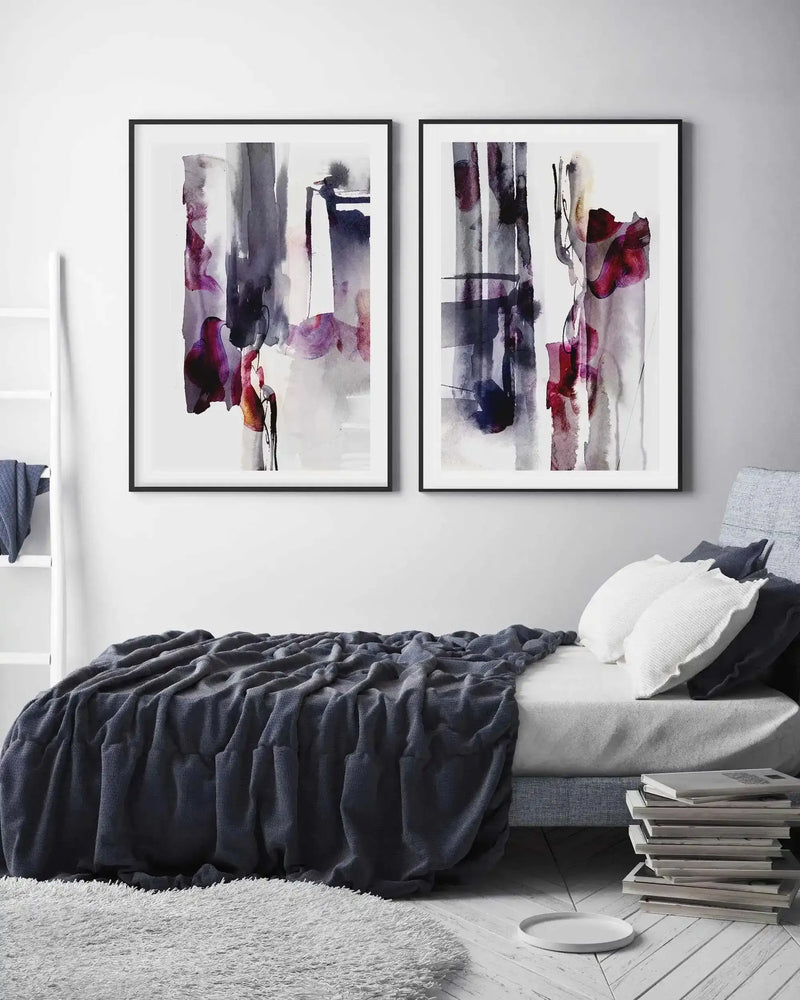 Two large framed abstract art prints displayed side-by-side above a bed in a minimalist bedroom. Both prints feature vertical brushstrokes in shades of deep charcoal, muted purple, and vibrant magenta, with black frames and white borders. The bed is covered with a dark grey textured throw, and a white fluffy rug is on the floor beside it.
