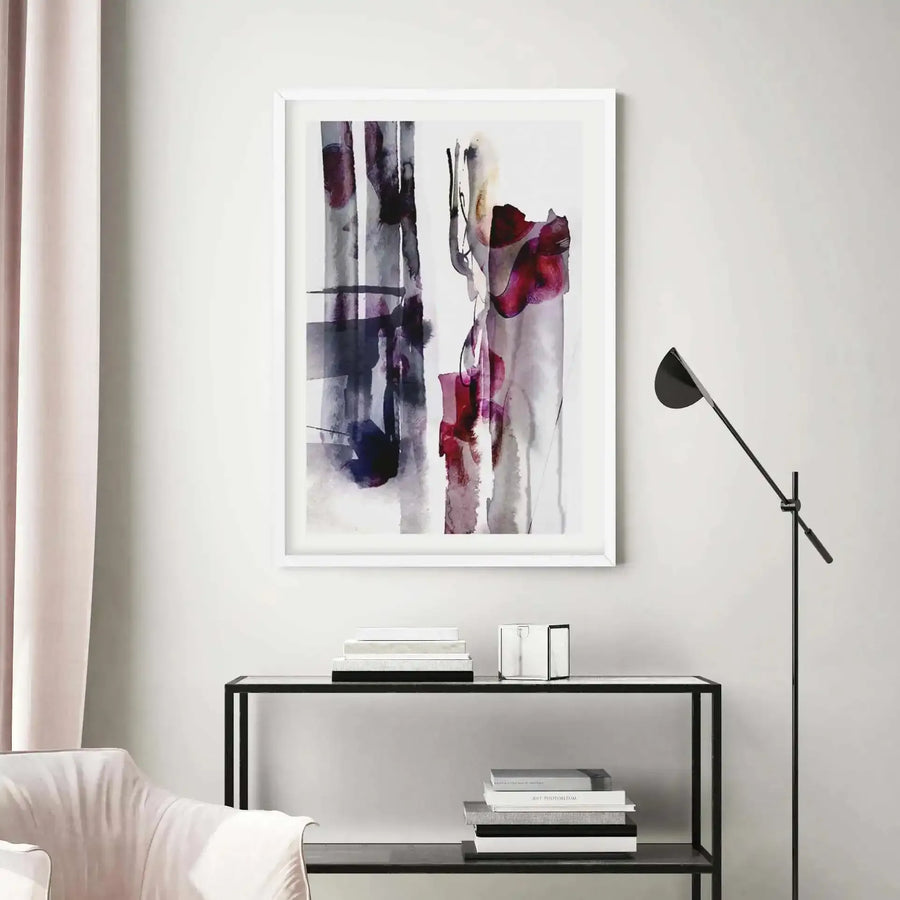 Large framed abstract art print displayed above a black console table in a minimalist living room. The print features vertical brushstrokes in shades of deep indigo, plum, and crimson, with a fluid, watercolour-like quality, set in a white frame with a white border. The room includes a pale pink curtain, a cream armchair, and a black floor lamp.