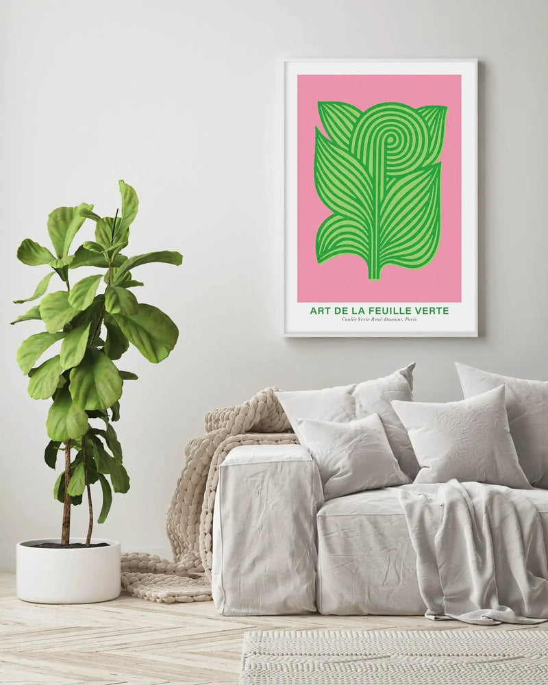 Large framed art print displayed above a white sofa in a minimalist living room. The print features an abstract illustration of a green leaf with swirling lines on a bright pink background, with a white frame and white border. The room includes a large fiddle leaf fig plant in a white pot, a chunky knit cream throw, and a light-coloured herringbone wood floor.