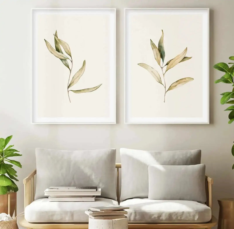 Two framed art prints displayed side-by-side above a light wood sofa with cream cushions in a minimalist living room. Each print features a single branch of Australian foliage with elongated, pale olive-green leaves and subtle brown undertones, rendered in a soft watercolour style on a cream background, with white frames and white borders. A potted green plant sits on either side of the sofa.