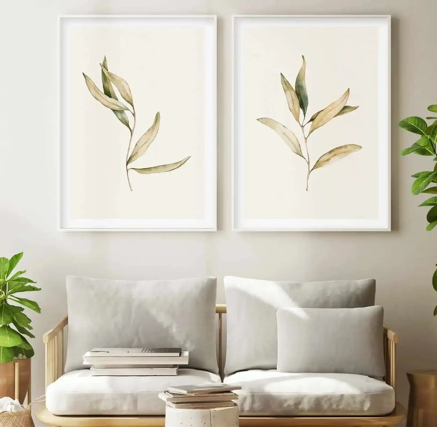 Two framed art prints displayed side-by-side above a light wood sofa with cream cushions in a minimalist living room. Each print features a single branch of Australian foliage with elongated, pale olive-green leaves and subtle brown undertones, rendered in a soft watercolour style on a cream background, with white frames and white borders. A potted green plant sits on either side of the sofa.