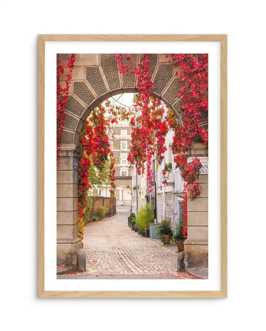 Framed photography art print of a cobblestone archway in London, with a natural wood frame and white border. The archway is covered in vibrant red and orange autumn ivy, leading to a narrow street lined with quaint buildings.
