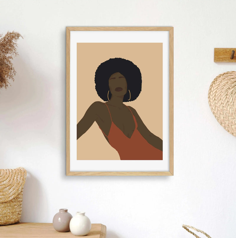 Framed art print displayed on a white wall above a wooden console table in a minimalist living room. The print features an illustration of a dark-skinned woman with a large afro, wearing a rust-orange slip dress and gold hoop earrings, against a warm beige background, with a natural wood frame and white border. The room includes dried pampas grass in a woven basket and two small ceramic vases on the table, and a woven straw fan hanging on the wall.