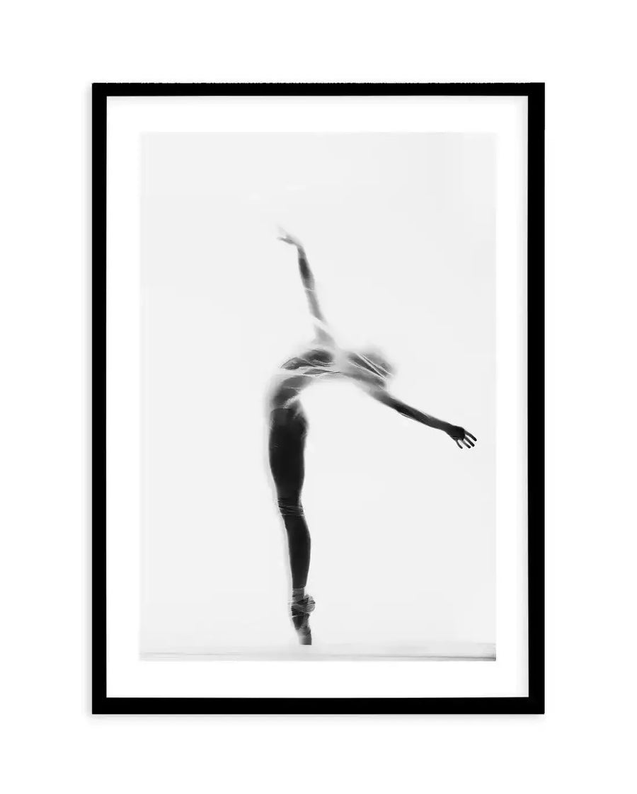 Framed black and white photograph of a ballerina en pointe, captured in silhouette behind a sheer, flowing fabric. The dancer is posed with her back arched, arms extended upwards and to the side, creating a graceful, ethereal form against a bright white background. The black frame has a white border.