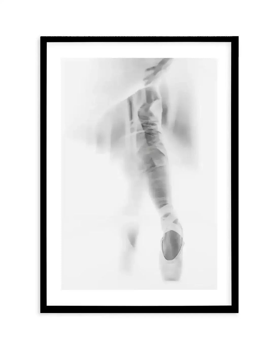 Framed black and white photograph of a ballerina en pointe, with a black frame and white border. The composition features the lower half of a dancer's body, with one leg extended and foot in a pointe shoe, creating a soft, blurred, and ethereal effect.