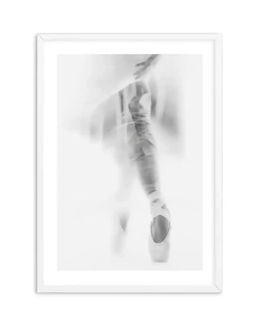 Framed black and white photograph of a ballerina en pointe, captured with a soft focus and motion blur effect. The composition features the lower half of a dancer, with one foot clearly visible in a pointe shoe, standing on the very tips of her toes. The image has a dreamlike, ethereal quality with blurred lines and subtle tonal variations from bright white to soft grey, displayed in a white frame with a white border.