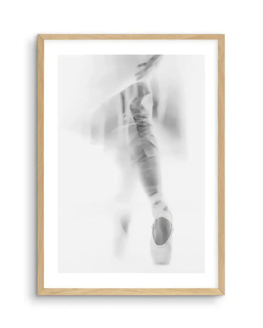 Framed black and white photograph of a ballerina en pointe, captured with a soft focus and motion blur effect, creating a dreamlike, ethereal quality. The dancer's leg and pointe shoe are visible, with the shoe appearing bright white against the blurred, lighter grey background, displayed in a natural oak frame with white border.