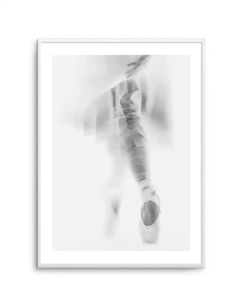 Framed black and white photograph of a ballerina en pointe, captured with a soft focus and motion blur effect. The composition features the lower half of the dancer's body, with one leg extended and foot in a pointe shoe, creating a graceful, ethereal silhouette against a bright white background. The print is displayed in a white frame with a white border.
