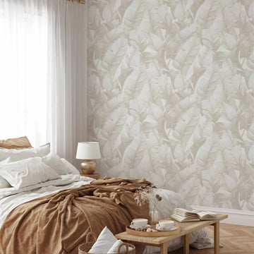 Lifestyle shot of a bedroom featuring a wall covered in "Banana Leaf Palms | Sepia Wallpaper". The wallpaper has a repeating pattern of large, overlapping banana leaves in muted sepia tones and warm cream, creating a soft, tropical feel. The room includes a bed with white bedding and a rich brown throw blanket, a gold-toned table lamp on a wooden bedside table, and a sheer white curtain covering a window. A small wooden bench at the foot of the bed holds a tray with two cups of coffee and a book.