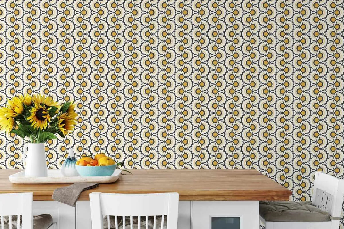 Bardot Retro Floral Wallpaper displayed on a kitchen wall behind a wooden countertop. The wallpaper features a repeating pattern of stylized sunflowers in golden yellow and olive green tones on a cream background with navy outlines. The design has a vintage 1970s aesthetic with geometric, slightly abstract flower motifs arranged in vertical columns. A white vase holding fresh sunflowers and a blue bowl with lemons sit on the counter, echoing the warm yellow tones of the wallpaper pattern.