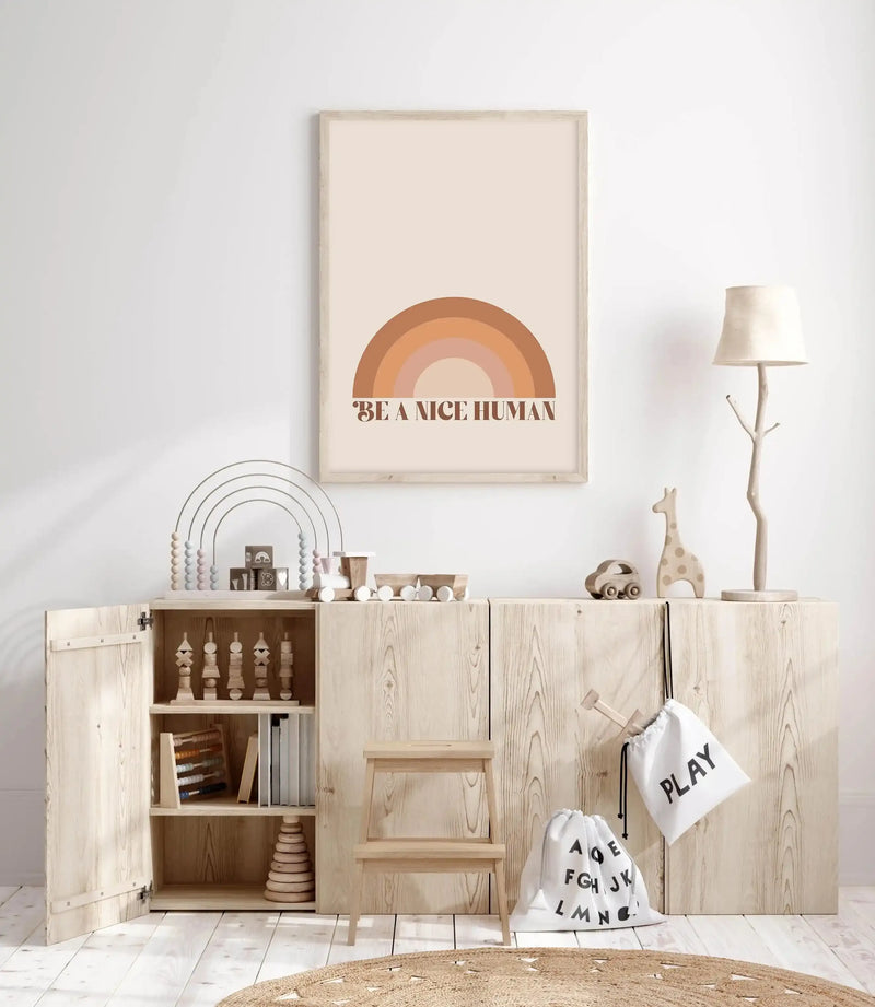 Kids Room:
Framed art print displayed above a wooden cabinet in a bright, minimalist kids' room. The print features a rainbow design in shades of brown, terracotta, and blush pink, with the text "BE A NICE HUMAN" in brown underneath, set against a cream background in a natural wood frame. The room includes wooden toys, a small wooden stool, and a woven rug.