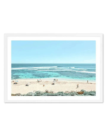 Framed aerial photograph of a pristine beach scene at Yallingup. The composition shows pale golden sand in the foreground with sparse coastal vegetation, a wide sandy beach with scattered beachgoers and umbrellas, turquoise and aquamarine shallow waters with white-capped waves, and a clear gradient sky transitioning from pale blue to deeper blue at the horizon. The photograph is printed on white paper and mounted in a white frame with a white border, displayed against a light grey outer frame.