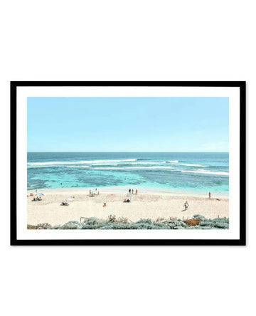 Framed aerial photograph displaying a coastal beach scene in Yallingup. The image shows a wide sandy beach with scattered beachgoers and surfboards, turquoise shallow water with white-capped waves, and darker ocean beyond under a clear pale blue sky. Native coastal vegetation in muted greens appears in the foreground. The photograph is mounted with a white border in a black frame.