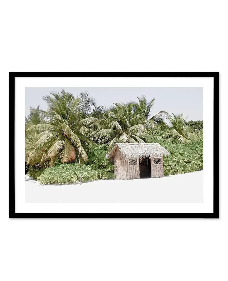 Framed photograph of a tropical beach scene with a small, rustic hut made of straw and wood on white sand, surrounded by lush green foliage and tall palm trees under a bright, clear sky, with a black frame and white border.