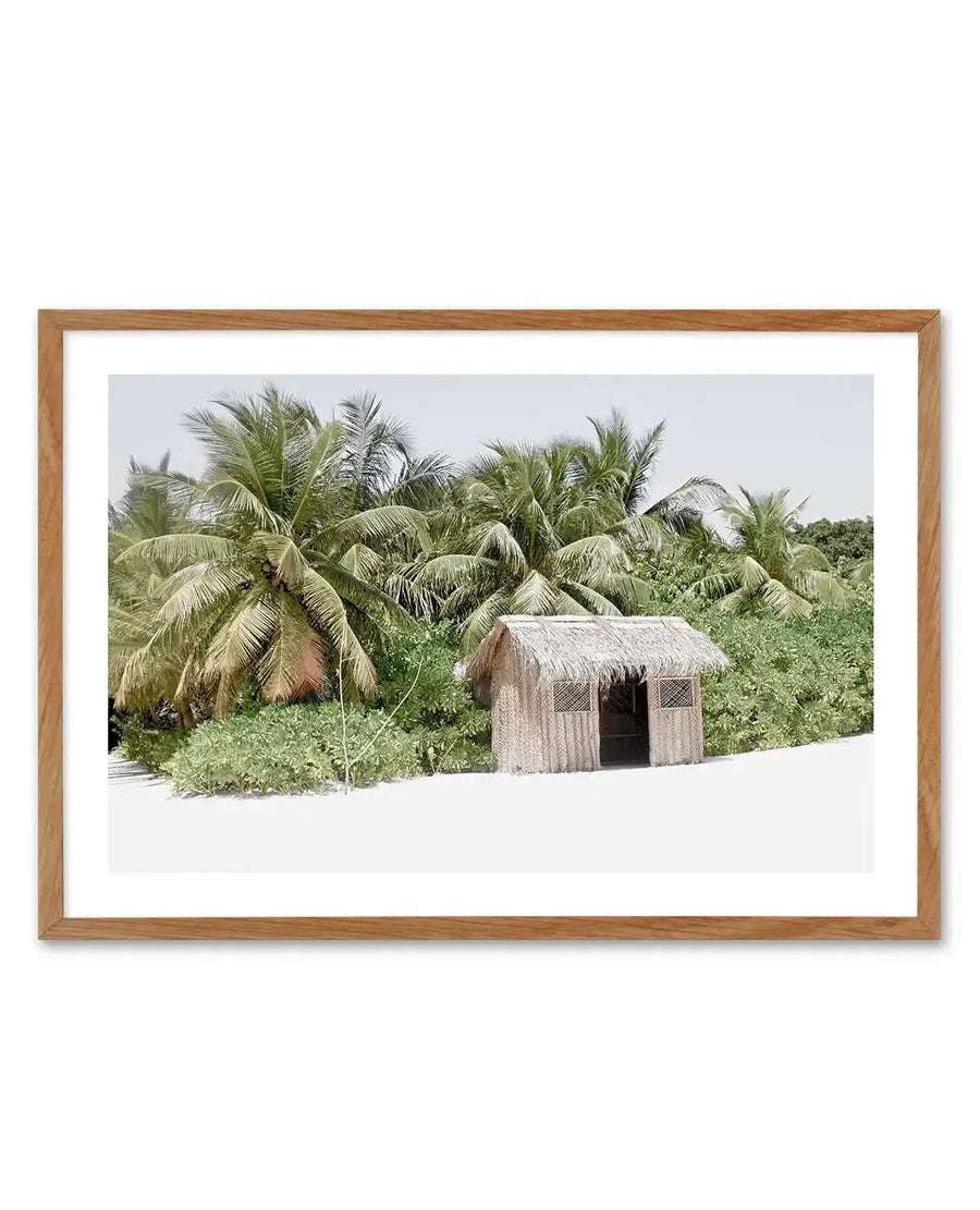 Framed photography art print of a tropical beach scene with a small, rustic hut, displayed in a natural oak frame with white border. The composition features a thatched-roof hut with two open windows and a doorway, nestled among lush green foliage and tall palm trees, with a bright white sandy foreground and a pale blue sky.