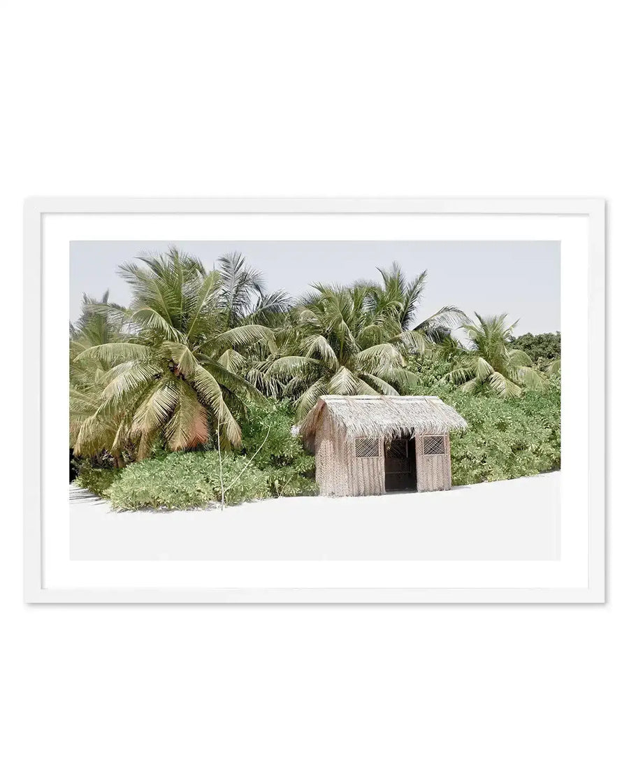 Framed photograph of a tropical beach scene with a white frame and white border. The image features a small, rustic beach hut with a thatched roof and wooden walls, nestled among lush green foliage and tall palm trees, with a bright white sandy foreground and a clear, light blue sky.