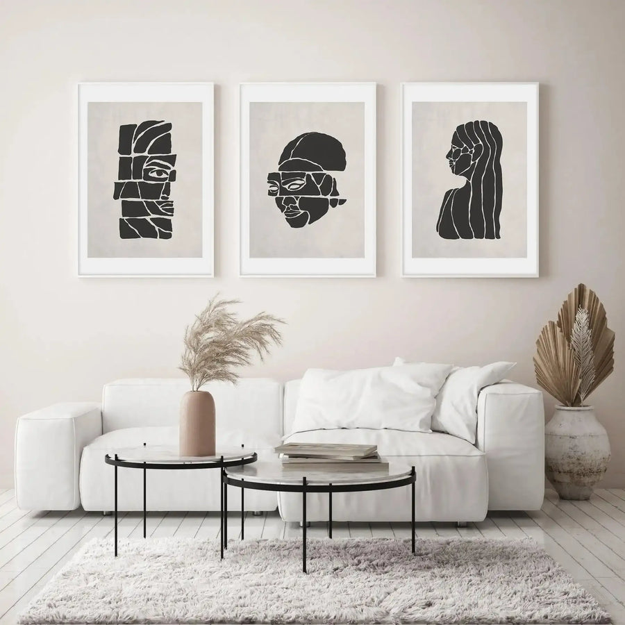 Three framed art prints displayed above a white sofa in a minimalist living room. The prints feature abstract black line art on a light cream background, with white frames and white borders. The left print shows a fragmented face, the middle print shows a fragmented face with a hat, and the right print shows a woman's profile with flowing hair. The room includes two round coffee tables with marble tops and black legs, a shaggy white rug, and dried pampas grass in a light brown vase.