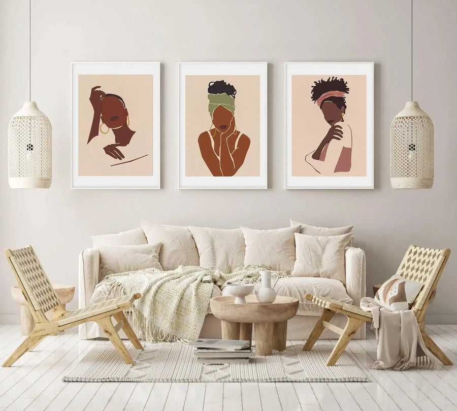 Lifestyle shot of three framed art prints displayed above a cream sofa in a minimalist living room. Each print features an abstract illustration of a Black woman's face and upper body in various poses, with dark brown skin tones and accents of muted green, dusty pink, and gold, against a light beige background, with white frames and white borders. The room includes two woven pendant lights, two woven wooden chairs, a round wooden coffee table, and a textured cream throw.