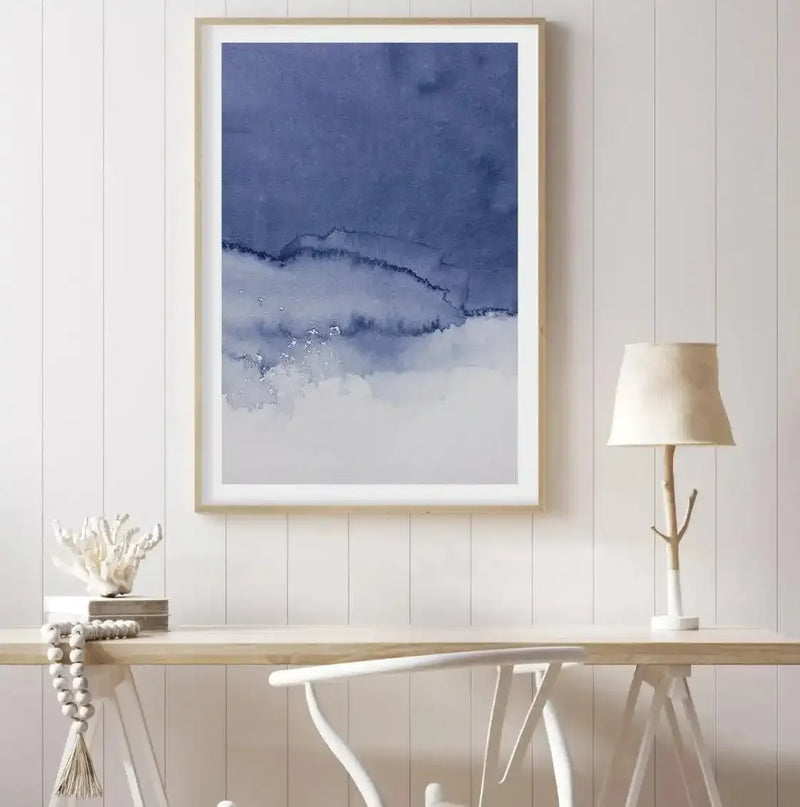 Home Office:
Large framed art print displayed above a light wood desk in a minimalist home office. The print features an abstract watercolor design with a deep indigo blue upper section transitioning into a lighter, more fluid blend of blues and whites, suggestive of ocean waves or a cloudy sky, with a natural wood frame and white border. The room includes a white chair, a white coral sculpture, a string of white wooden beads, and a table lamp with a natural branch base and a cream fabric shade.