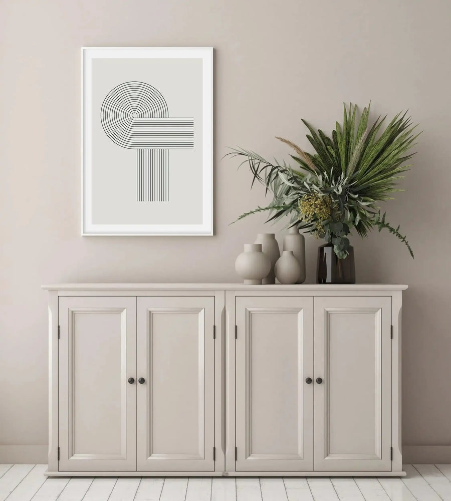 Lifestyle shot featuring a framed art print displayed above a cream-coloured wooden cabinet in a minimalist room with light beige walls and white wooden floorboards. The art print features a geometric design of concentric curved and straight lines in black on a light grey background, with a white frame and white border. A large vase of dried green and brown foliage sits on the cabinet to the right of the print, surrounded by smaller cream-coloured vases.