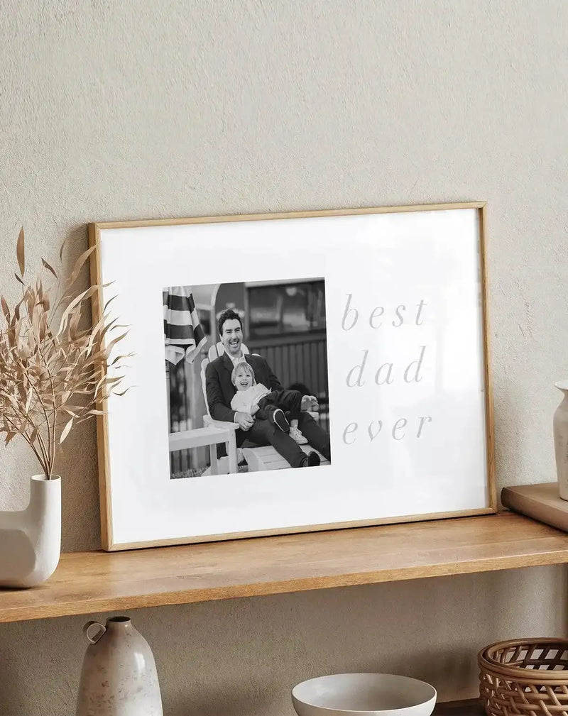 Framed art print with natural wood frame displaying a black and white photograph of a man holding a young child, positioned on the left side of white background with gray cursive text reading "best dad ever" on the right. White border surrounds the design. Print sits on wooden shelf next to cream ceramic vase with dried pampas grass, small ceramic vessel, and woven basket. Textured beige wall visible in background.
