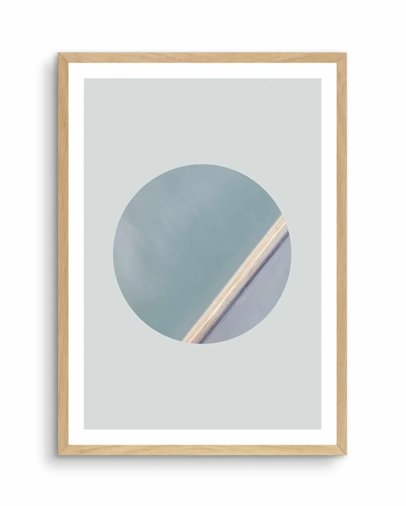 Framed art print featuring a minimalist composition with a large muted teal-blue circle on a pale grey background. A diagonal stripe in cream and soft purple tones cuts through the lower right portion of the circle. The artwork is displayed in a light natural wood frame with a white border, mounted on a white wall.