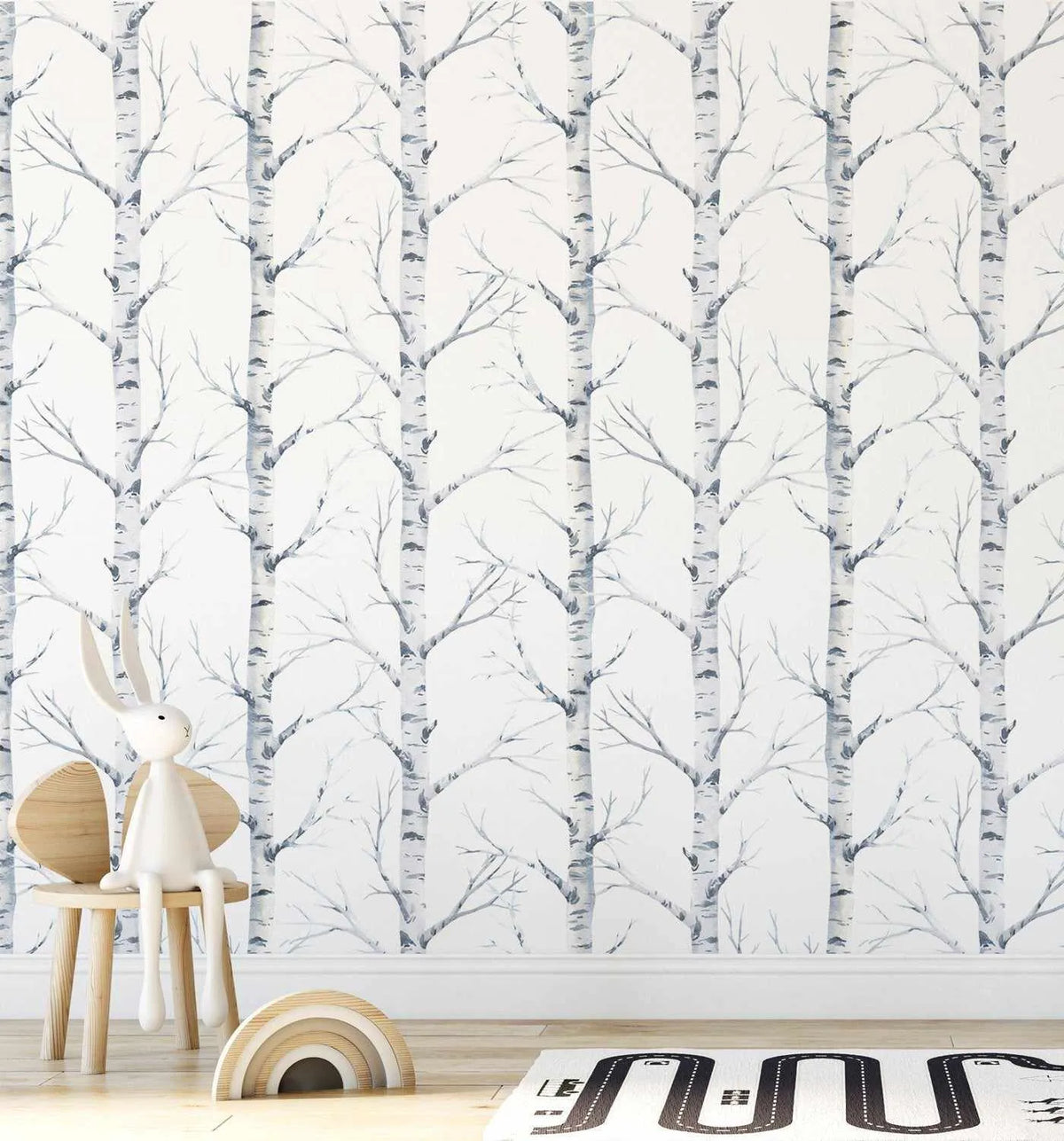 Nursery:
"Peel and stick wallpaper featuring a repeating pattern of hand-painted birch trees with white trunks and delicate bare branches in shades of grey and white on a soft white background. The wallpaper is displayed in a child's room with a small wooden chair holding a white rabbit figurine, a wooden rainbow toy, and a black and white road map rug on a light wood floor."