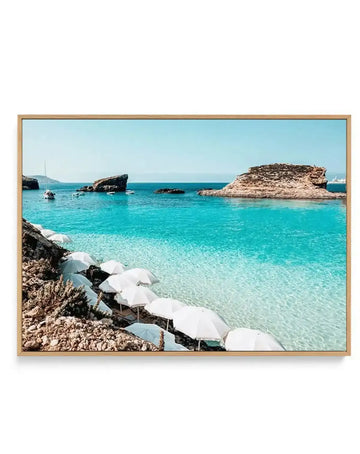 Framed horizontal canvas print featuring a Mediterranean beach scene in Blue Lagoon, Malta. The photograph shows crystal-clear turquoise water with rocky outcrops and small islands, a row of white beach umbrellas along the foreground rocks, and sailboats anchored in the shallow bay. The sky is pale blue and cloudless. The canvas is mounted in a natural wood frame with a light tan finish.