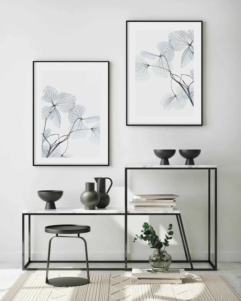 Lifestyle shot of a minimalist living room featuring two framed art prints with black frames and white borders, displayed on a white wall above a console table. The prints feature delicate, skeletal blue leaves with intricate vein patterns against a bright white background. The console table is black with a white marble top, adorned with black ceramic bowls, vases, and a small plant in a glass vase. A black metal stool and a textured cream rug are also visible.