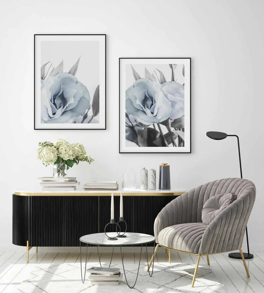 Lifestyle shot of a modern living room featuring two framed art prints above a black console table. Each print displays a close-up photograph of a light blue lisianthus flower with soft, delicate petals and grey-green leaves, set against a bright white background, with a black frame and white border. The room includes a grey ribbed armchair, a marble coffee table with candles, and a vase of white hydrangeas.