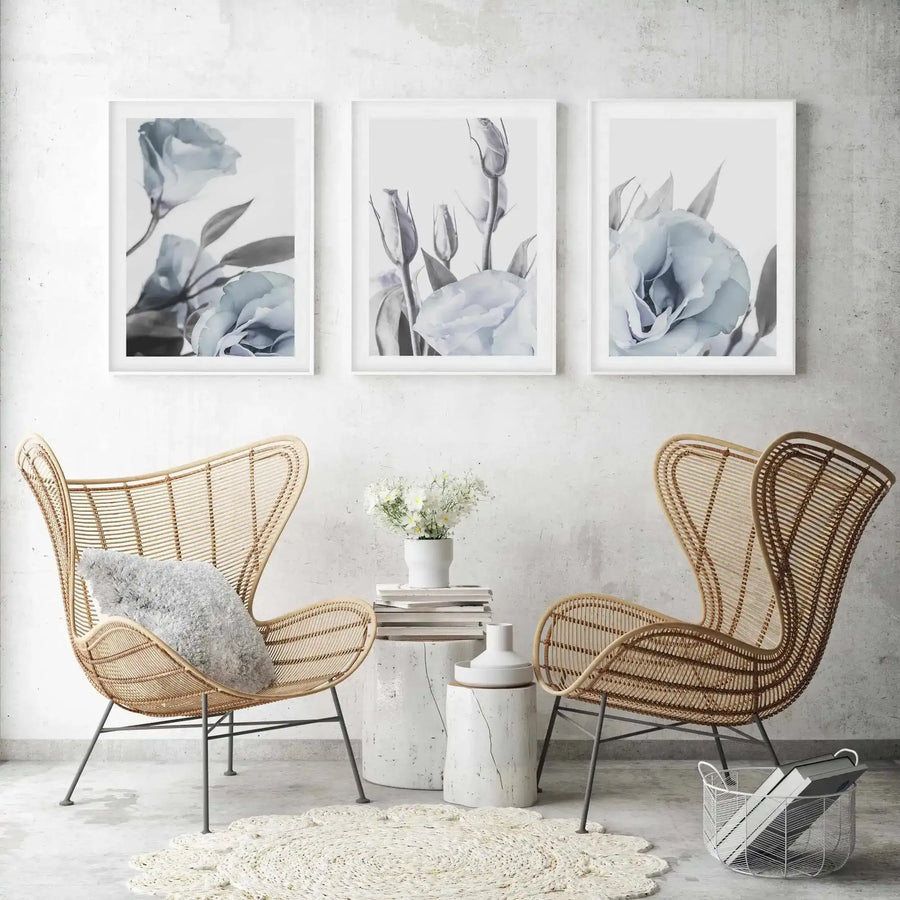 Three framed art prints displayed above two rattan chairs in a minimalist living room. Each print features a blue lisianthus flower or buds with grey-green leaves on a white background, with a white frame and white border. The room includes two white ceramic side tables, a white vase with white flowers, a white woven rug, and a white wire basket with books.