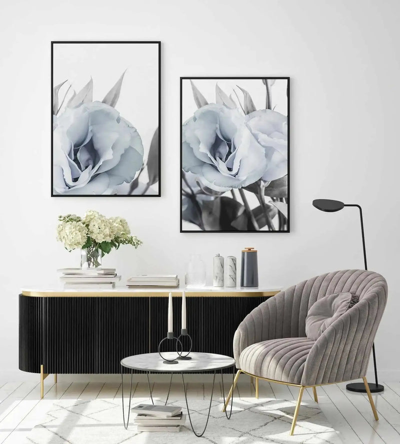 Lifestyle shot of two framed canvas art prints displayed on a white wall above a black fluted console table in a modern living room. Each print features a close-up black and white photograph of a delicate blue lisianthus flower with soft, layered petals and visible stems, framed in black with a white border. The room includes a grey velvet armchair, a round marble coffee table with candles, and a vase of white hydrangeas.