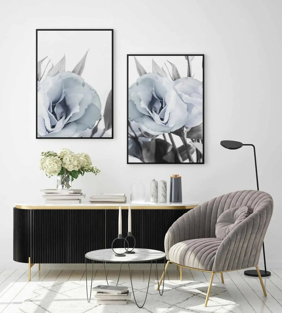 Lifestyle shot of two framed canvas art prints displayed on a white wall above a black fluted console table in a modern living room. Each print features a close-up black and white photograph of a delicate blue lisianthus flower with soft, layered petals and visible stems, framed in black with a white border. The room includes a grey velvet armchair, a round marble coffee table with candles, and a vase of white hydrangeas.