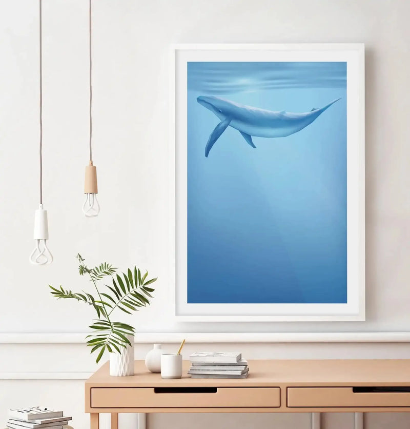 Home Office:
Large framed art print featuring a blue whale swimming underwater, displayed above a light wood desk in a minimalist home office. The print depicts a light blue whale with subtle shading, seen from below, against a gradient of light to deep blue water with sun rays filtering from the surface. The room includes two pendant lights with exposed filament bulbs, a green leafy plant in a white textured vase, and a stack of books on the desk.