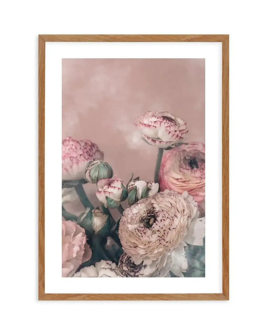 Framed art print of blush pink ranunculus flowers with layered petals against a soft mauve background. The composition features several blooms in varying stages of opening, with visible green stems and buds. Muted pink, cream, and white tones throughout. Natural photographic style with soft lighting and subtle shadows. Displayed in a natural wood frame with white border.