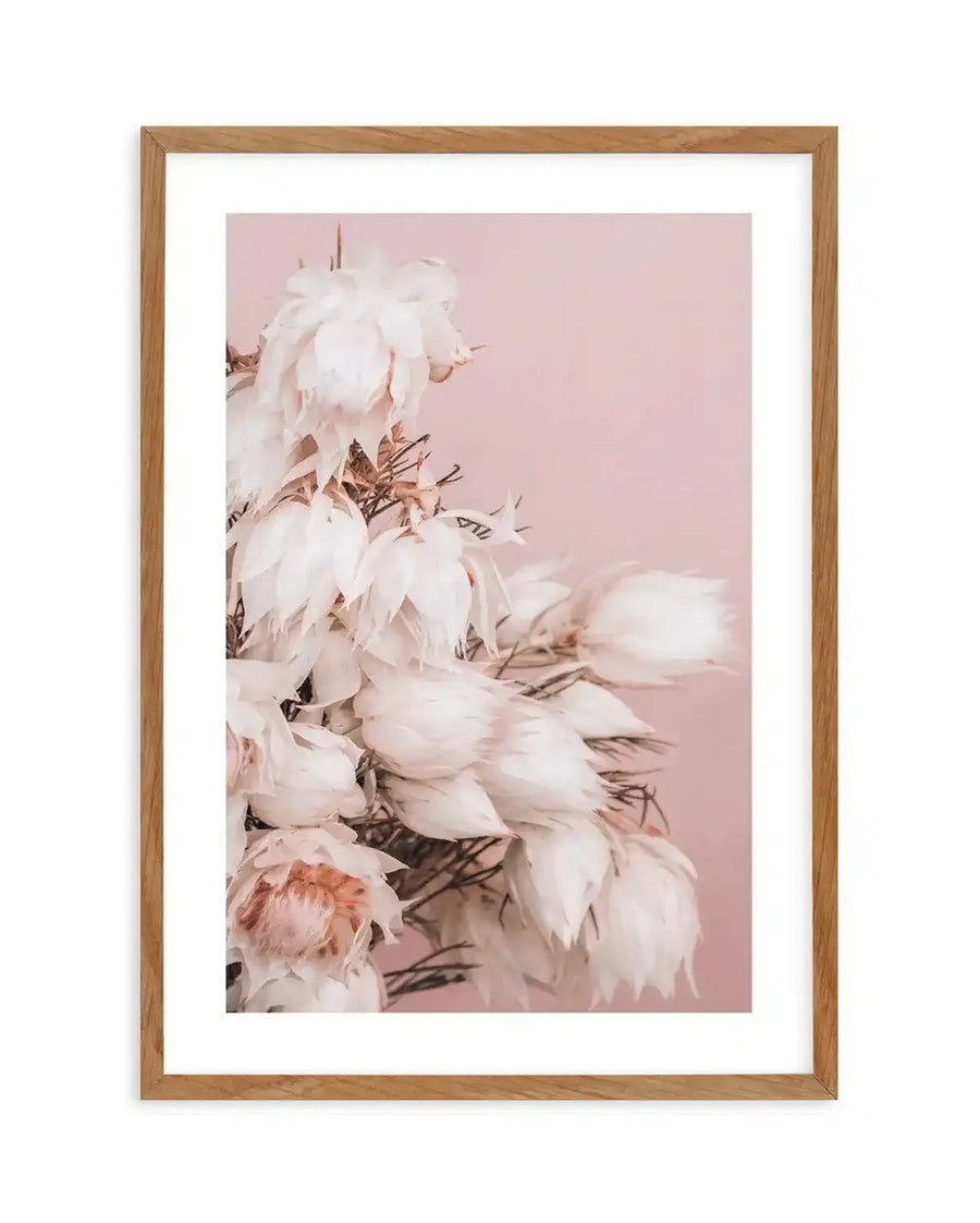 Framed art print featuring a soft focus photograph of white and blush pink peonies against a dusty rose background. The flowers are arranged vertically on the left side with delicate green stems and foliage visible throughout. The print is mounted in a natural wood frame with a white border, creating a soft, romantic aesthetic with muted, pastel tones throughout the composition.