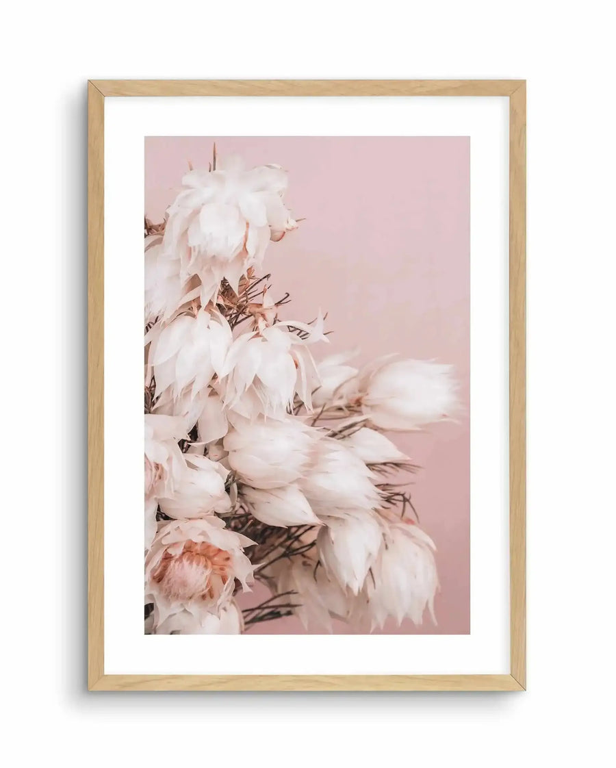Framed art print of delicate white and pale pink flowers with brown stems and stamens against a soft dusty rose background. Natural wood frame with white border surrounding the vertical portrait-orientation photograph. The flowers appear soft and slightly wilted, creating a romantic, muted aesthetic with warm neutral tones throughout the composition.