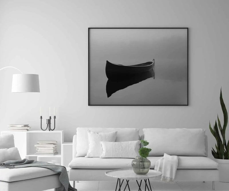 Black and white photograph of a solitary boat tilted at an angle against a neutral gray background, displayed in a black frame on a light gray wall above a white sofa in a minimalist living room setting.