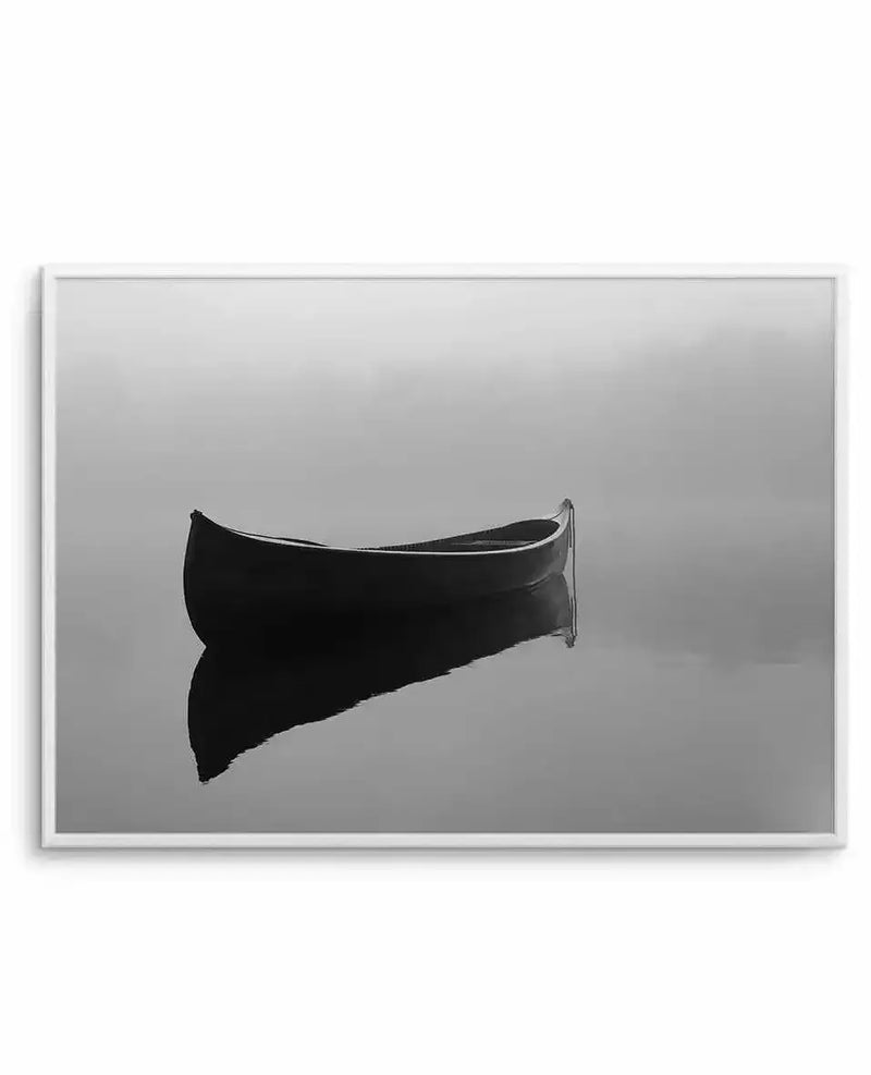 Black and white photograph of a solitary dark boat on still water, shown in profile against a misty, overcast gray sky. The boat appears as a dark silhouette with subtle reflections visible on the calm water surface. The minimalist composition creates a serene, contemplative mood. The artwork is presented in a white-bordered horizontal frame.